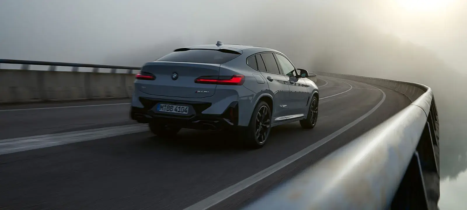 BMW X4 رمادية من الخلف أثناء القيادة على جسر منحنٍ وسط الضباب