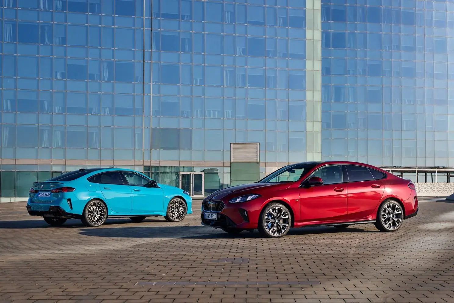 سيارتا BMW سيدان زرقاء وحمراء أمام مبنى زجاجي، الحمراء جانبية من الأمام والزرقاء من الخلف