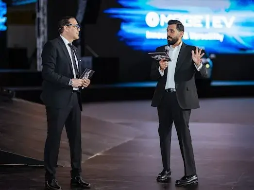 متحدثان على المسرح خلال حفل إطلاق SN Automotive وخلفهما شاشة سيارة MAGE
