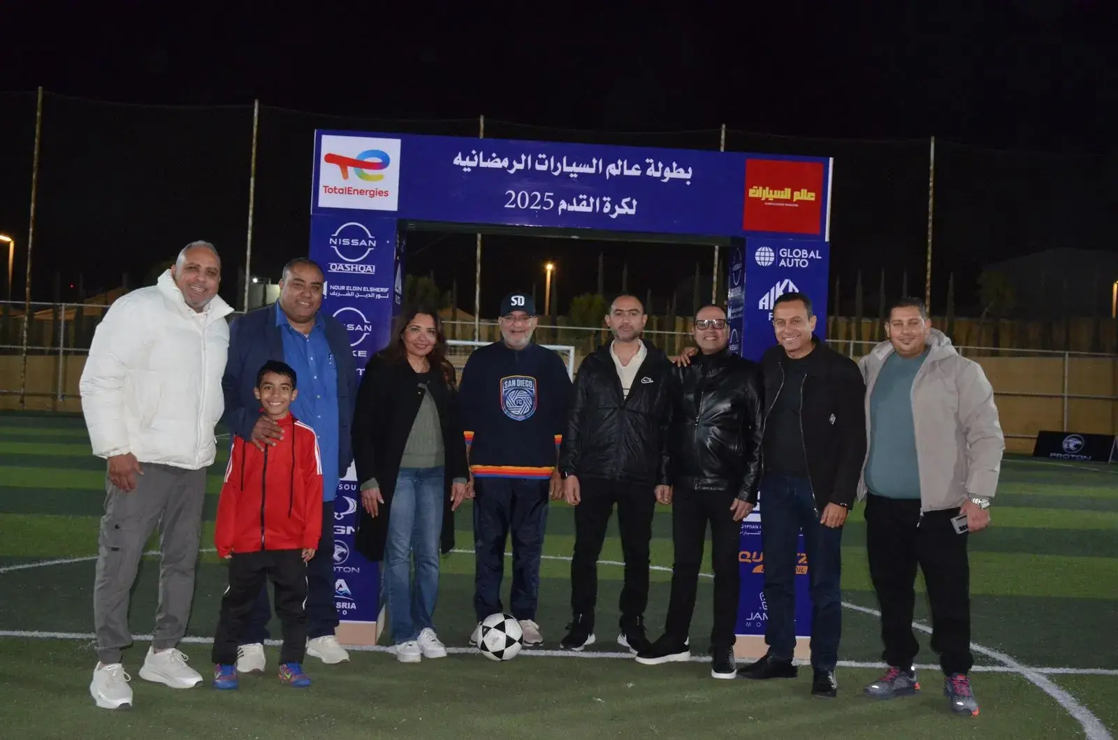 مجموعة من المشاركين يقفون أمام بوابة البطولة الرمضانية لكرة القدم على ملعب عشبي ليلًا